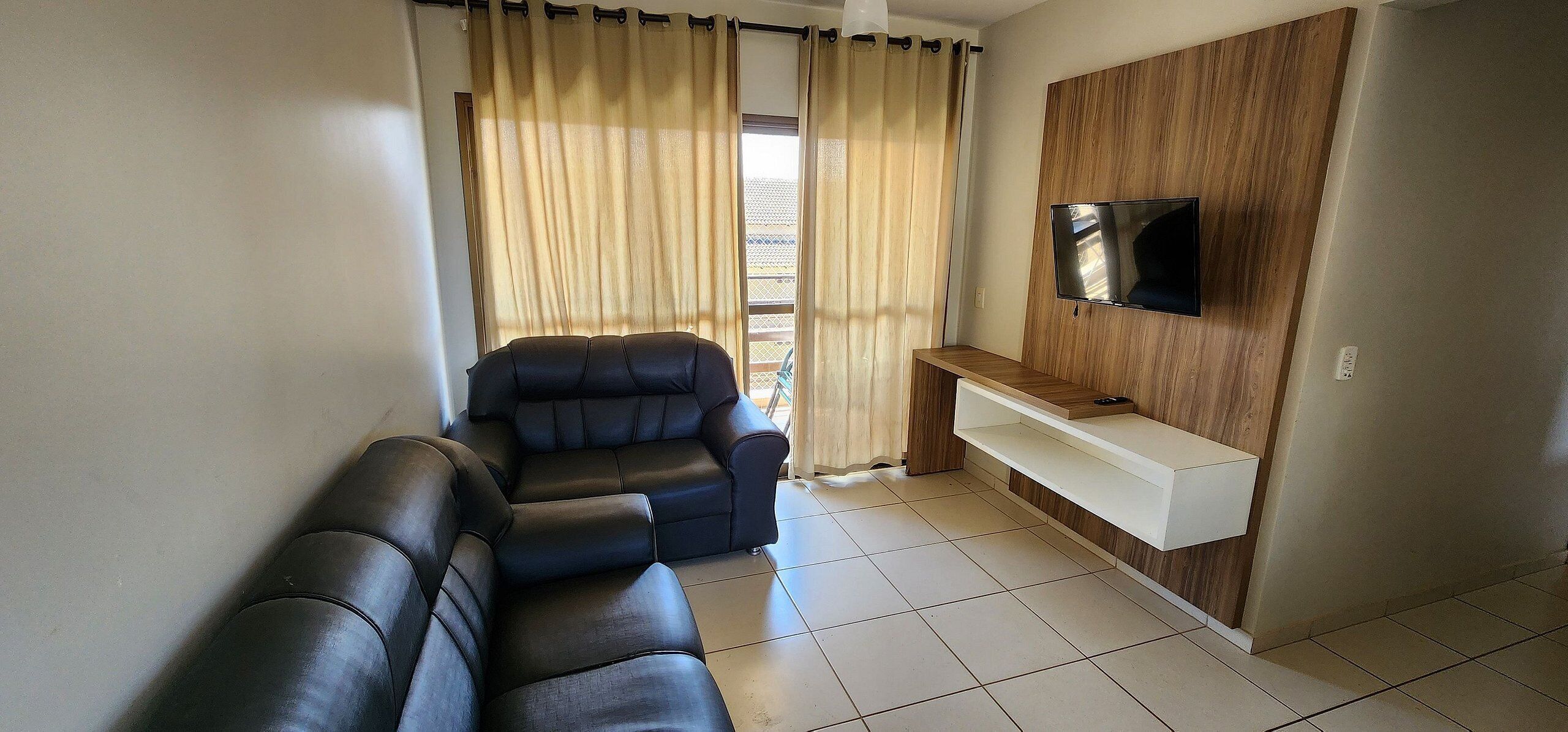 A living room with a sofa, a recliner, a TV mounted on a wooden wall, and a window with curtains.