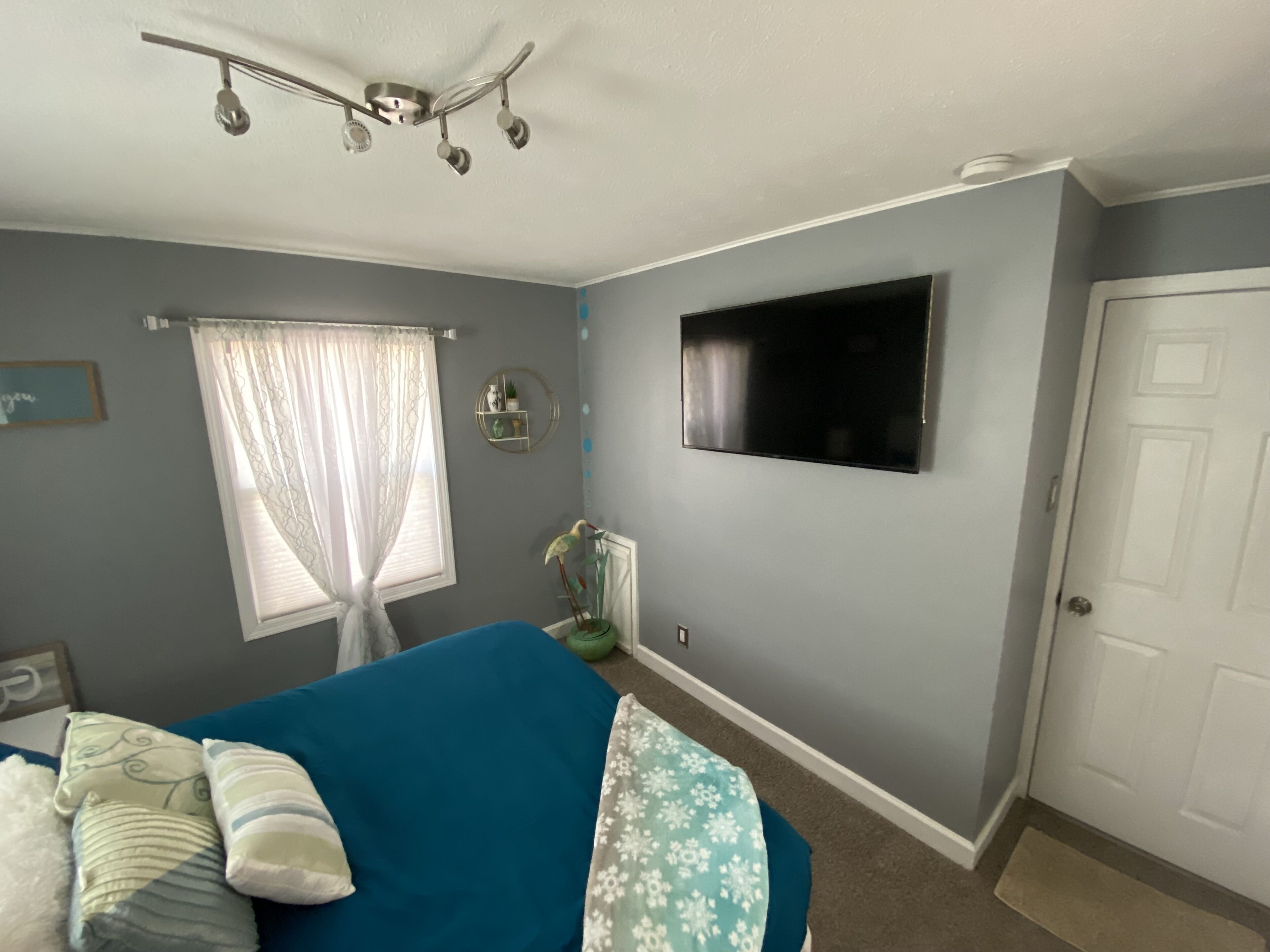 Un dormitorio con cama, televisor montado en la pared, ventana con cortinas y una puerta.