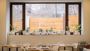 A dining area with tables set for two, potted plants on a window sill, and a view of a wooden fence outside. - Taniqinn Dilijan Hotel (Dilijan)