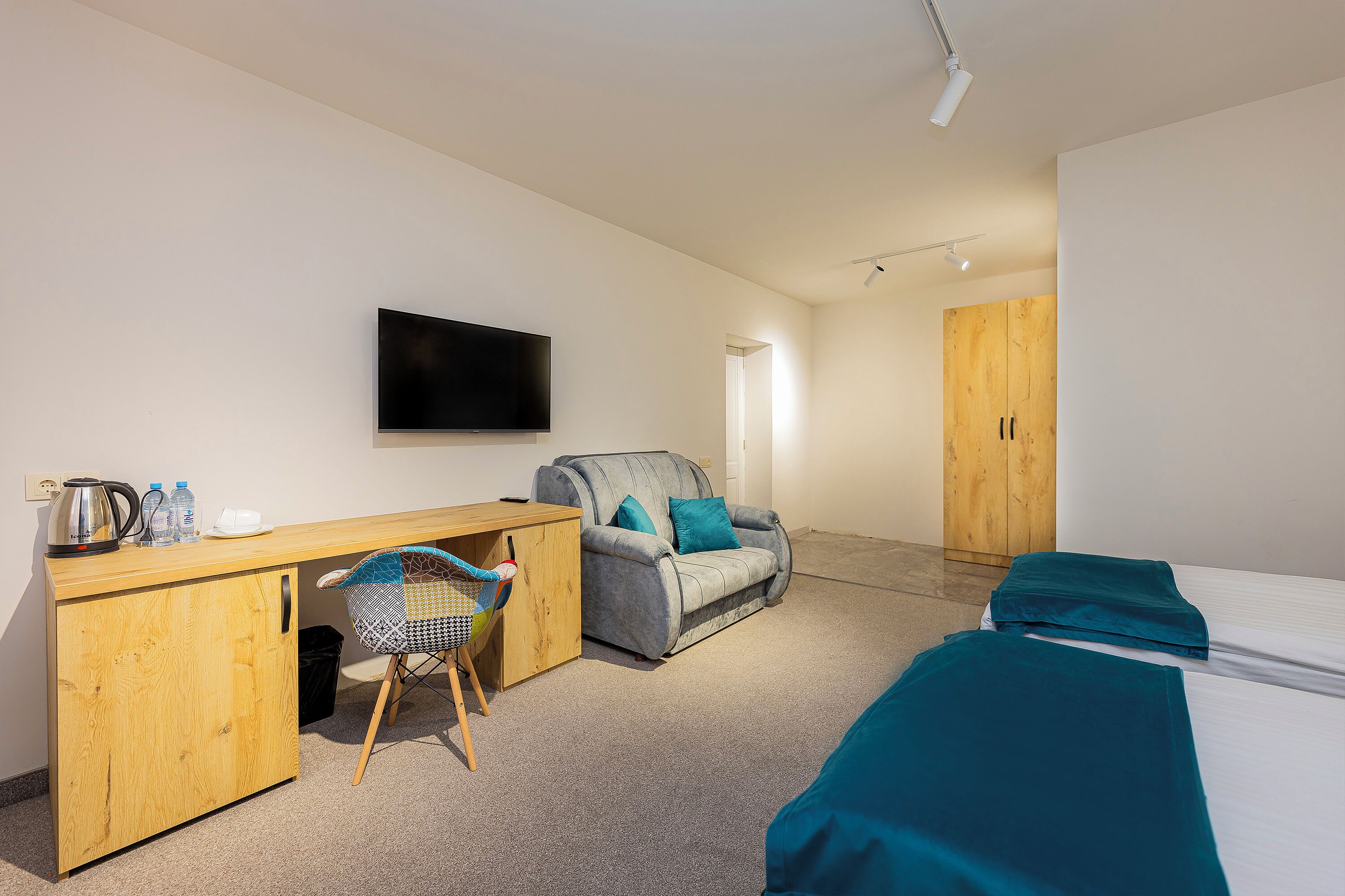 A hotel room with a wooden desk, a grey sofa, a TV mounted on the wall, and a bed with teal bedding.