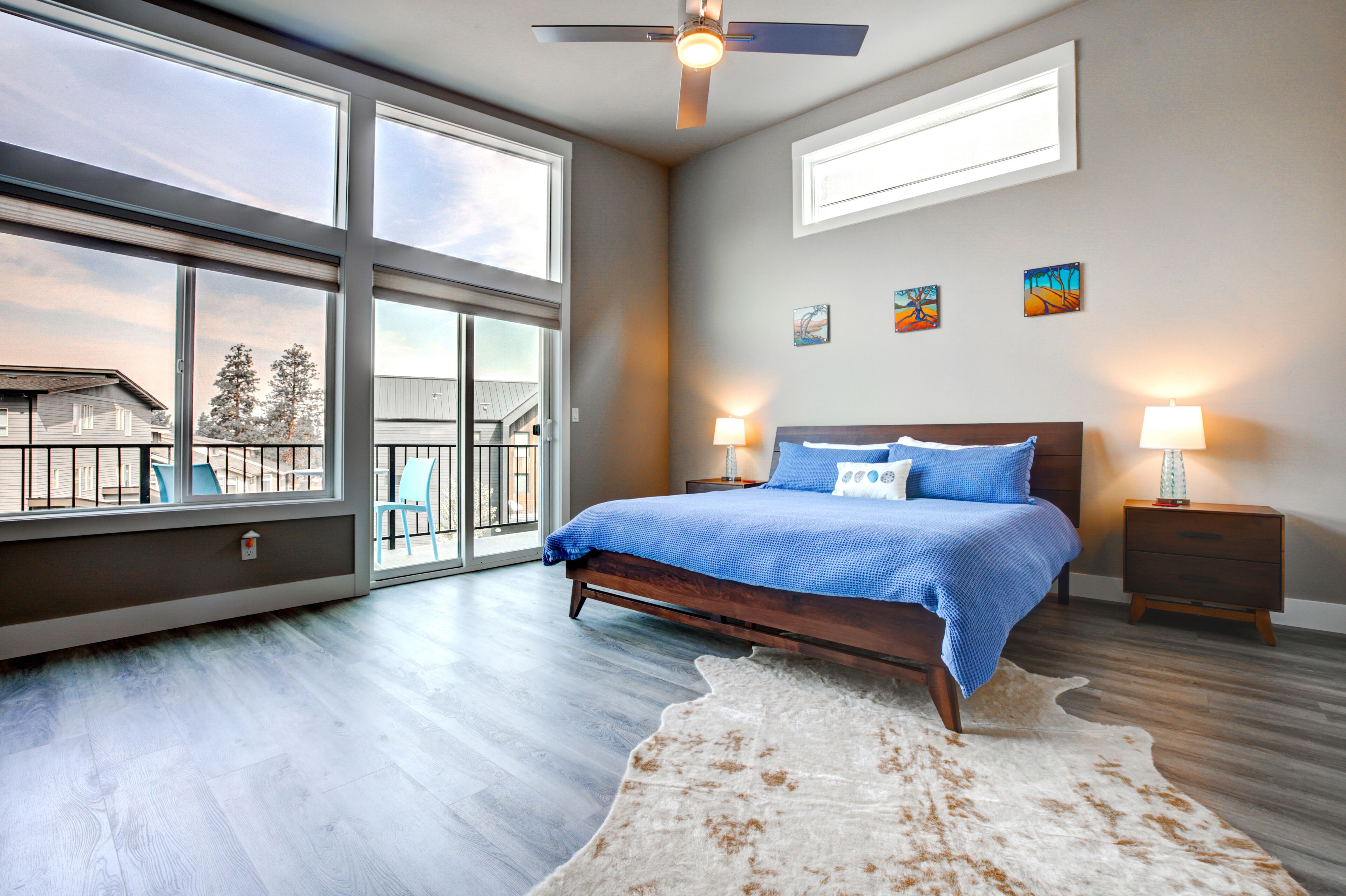 A bedroom with a large window, a ceiling fan, a bed with blue bedding, two bedside tables with lamps, and a cowhide rug.