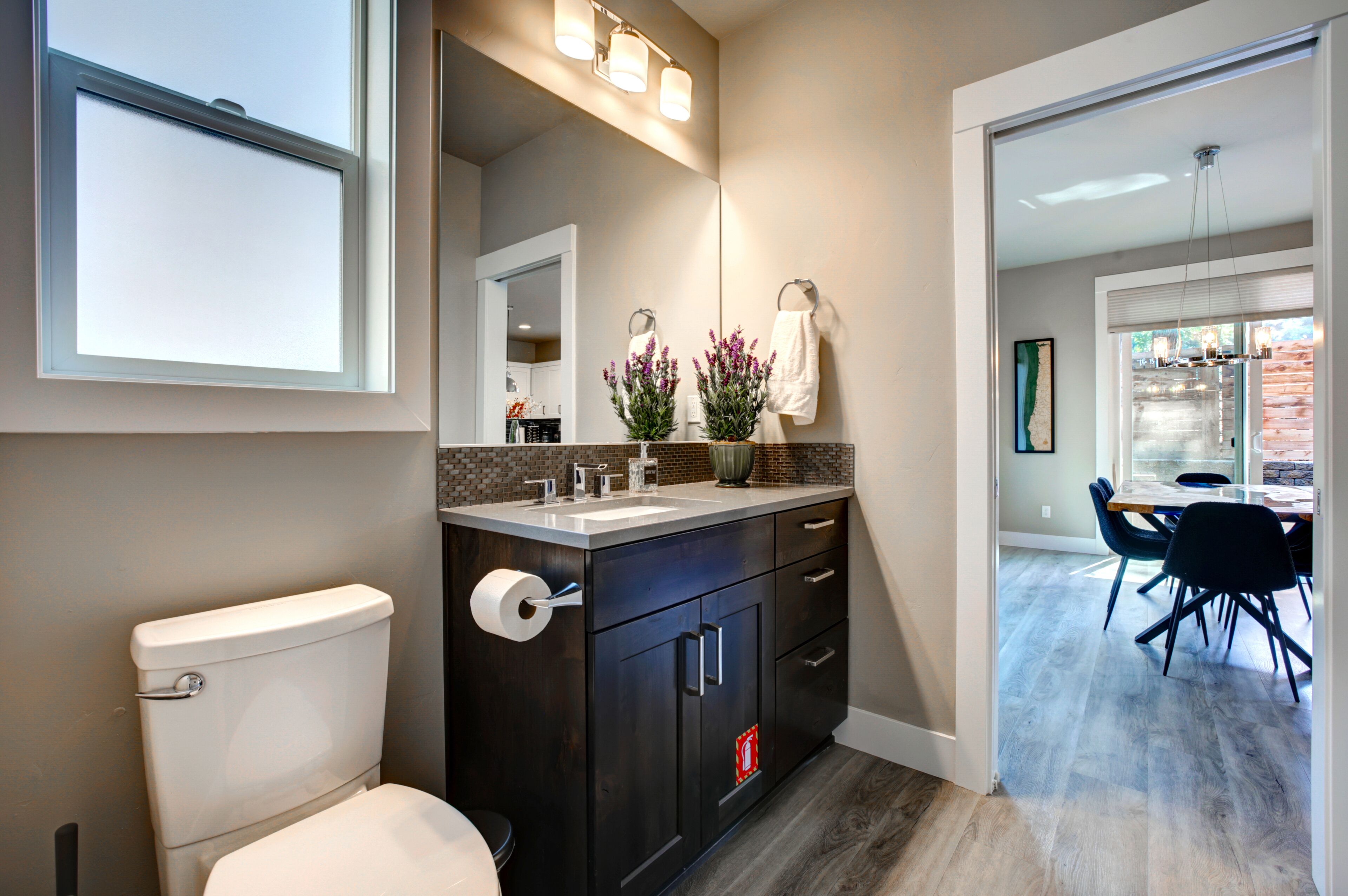 A modern bathroom with a toilet, dark wood vanity, a large mirror, and a window.