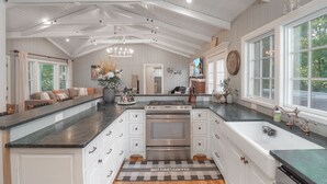 A spacious kitchen with a large island, white cabinetry, and a view of the outdoors through large windows. - Luxury Riverfront Escape- Hot tub & Fire Pit (Roseburg)