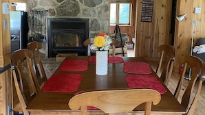 A wooden dining table with red placemats, surrounded by wooden chairs with cushioned seats. A stone fireplace with a black mantel is visible in the background. - Superbe Chalet au Coeur de la Nature et à Proximité D'un Corridor Aérobique (Montcalm)
