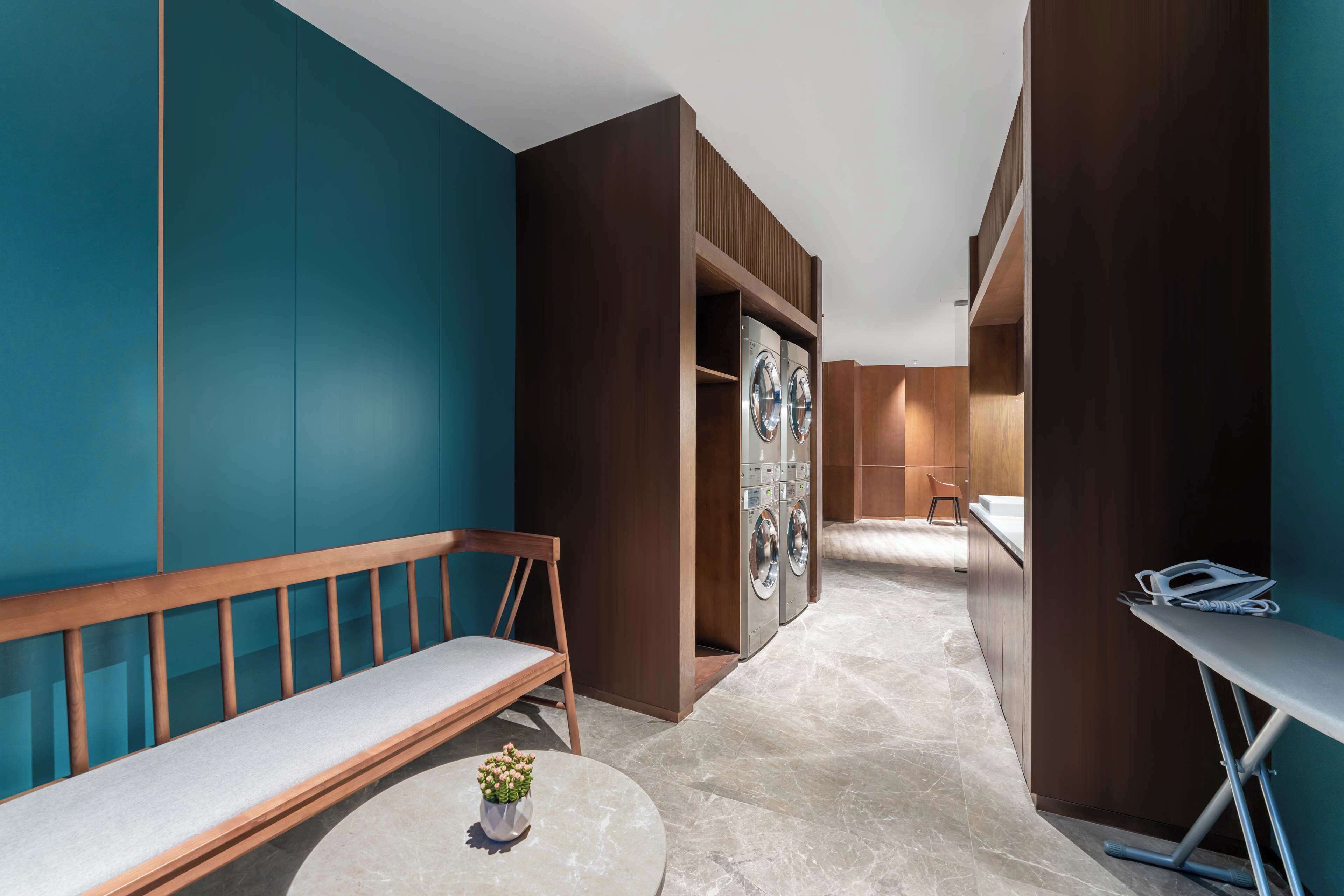 Una habitación de hotel moderna con un banco, una mesa redonda con un florero, una lavadora y una tabla de planchar pequeña.