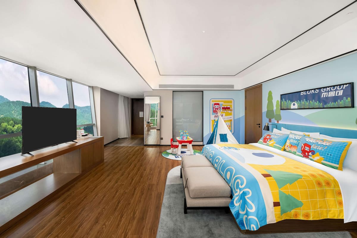 Una habitación de hotel moderna con un televisor grande, una cama con ropa de cama colorida, un escritorio con computadora y vistas a las montañas a través de ventanas amplias.