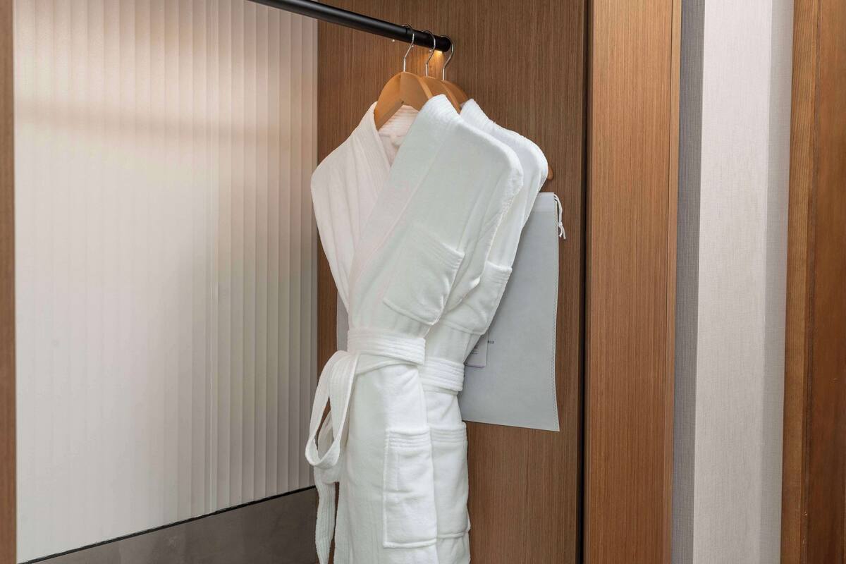 Un albornoz blanco colgado en un perchero de madera en una habitación de hotel con un panel de pared de madera y una ventana con persianas verticales.