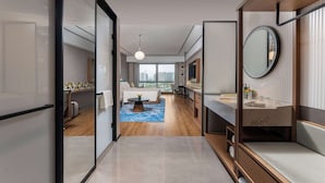 Ein modernes Hotelzimmer mit einem großen Bett, einer Sitzecke mit Couchtisch, einem Badezimmer mit Waschtisch und Spiegel sowie Blick auf die Stadt durch große Fenster.