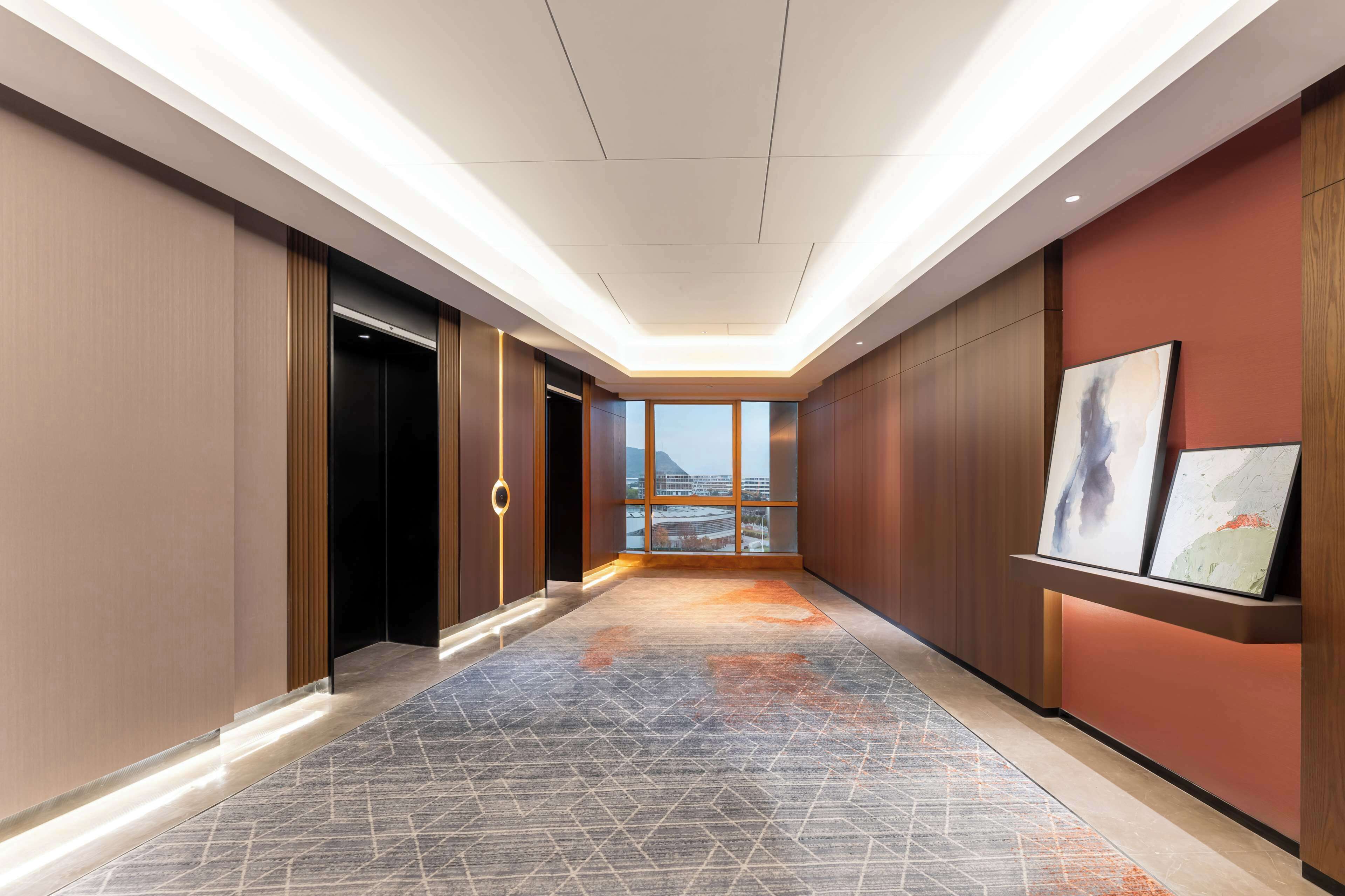 A hotel corridor with a geometric patterned floor, wooden panel walls, and a view of a cityscape through large windows.