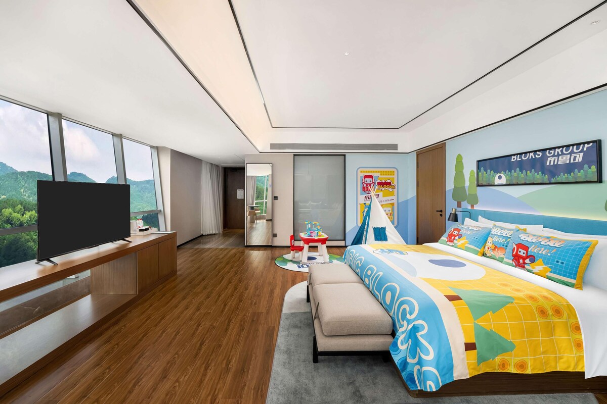 Ein modernes Hotelzimmer mit großem Fernseher, einem Bett mit bunter Bettwäsche, einem Schreibtisch mit Computer und Bergblick durch große Fenster.