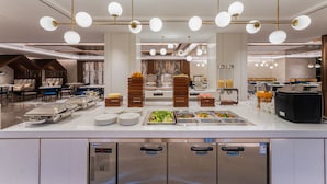 Ein modernes Buffet mit mehreren Speiseausgaben, einem Sitzbereich mit Tischen und Stühlen sowie einer Küche im Hintergrund.