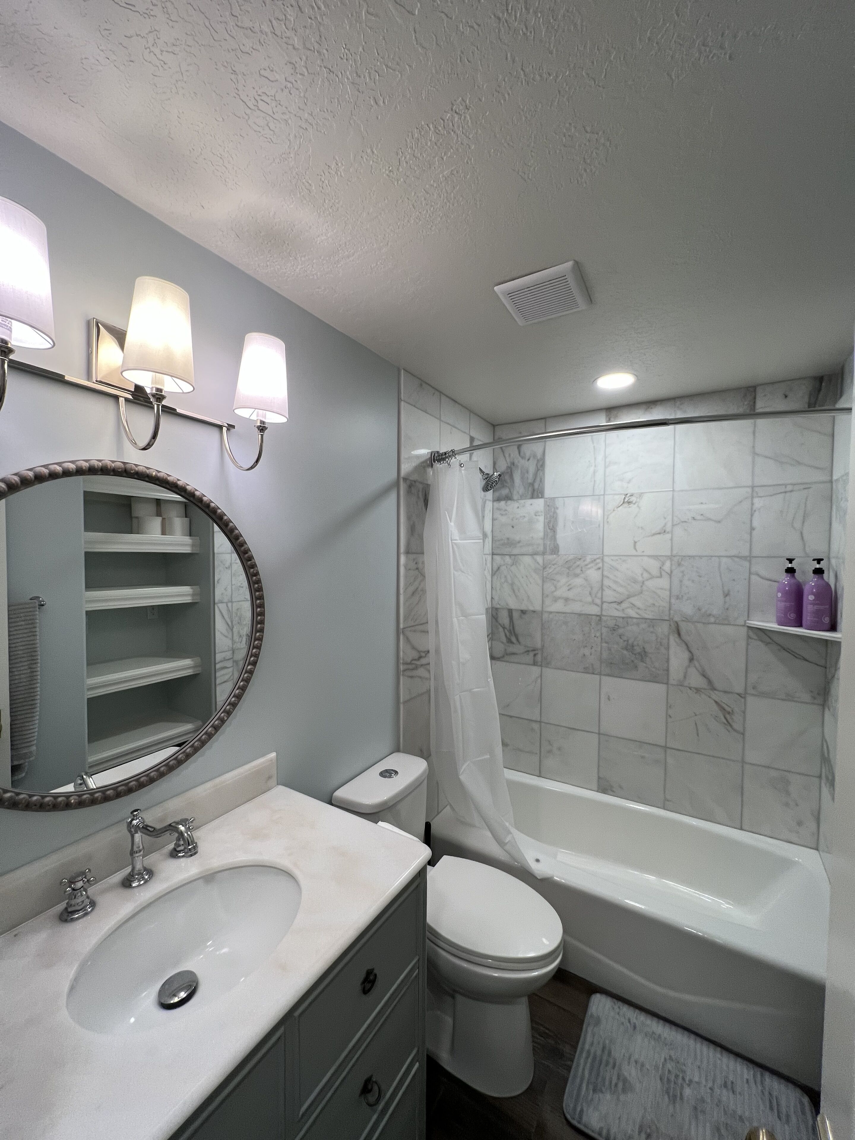 Un baño con lavamanos, inodoro y bañera. Hay repisas con artículos de tocador y un espejo con luces.