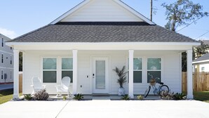 Ein eingeschossiges Haus mit Giebeldach, einer Veranda mit zwei weißen Stühlen, einem Fahrrad und einem Blumentopf.