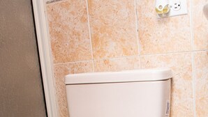 En toalett med ett vitt toalettlock och en vit cistern, en grå papperskorg och ett vägguttag med ett stickkontakt.