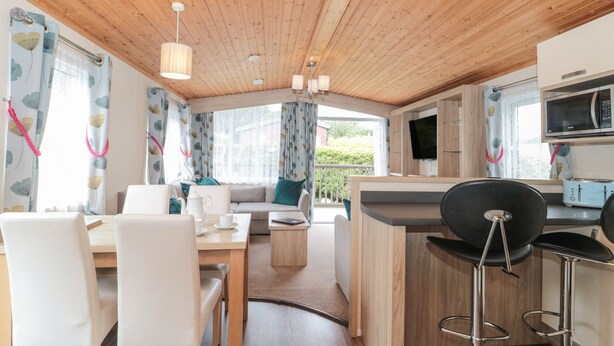 A cozy cabin-style room with a wooden ceiling, a dining area with a table and chairs, a sofa, a kitchen area with a microwave and toaster, and a large window with curtains. - Beech (Swanage)