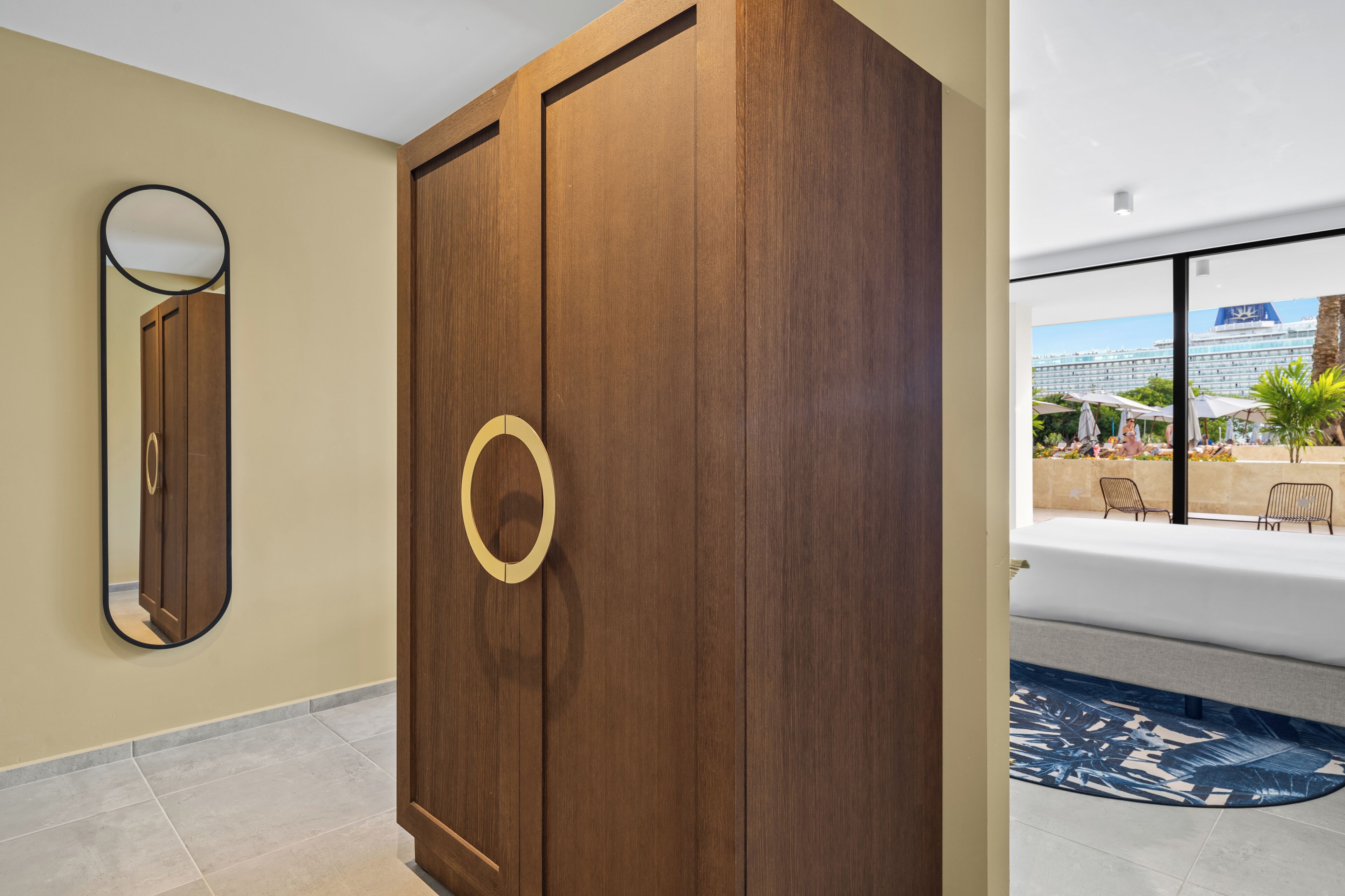 A wooden wardrobe with a circular handle, a mirror, and a view of a pool area with outdoor seating.