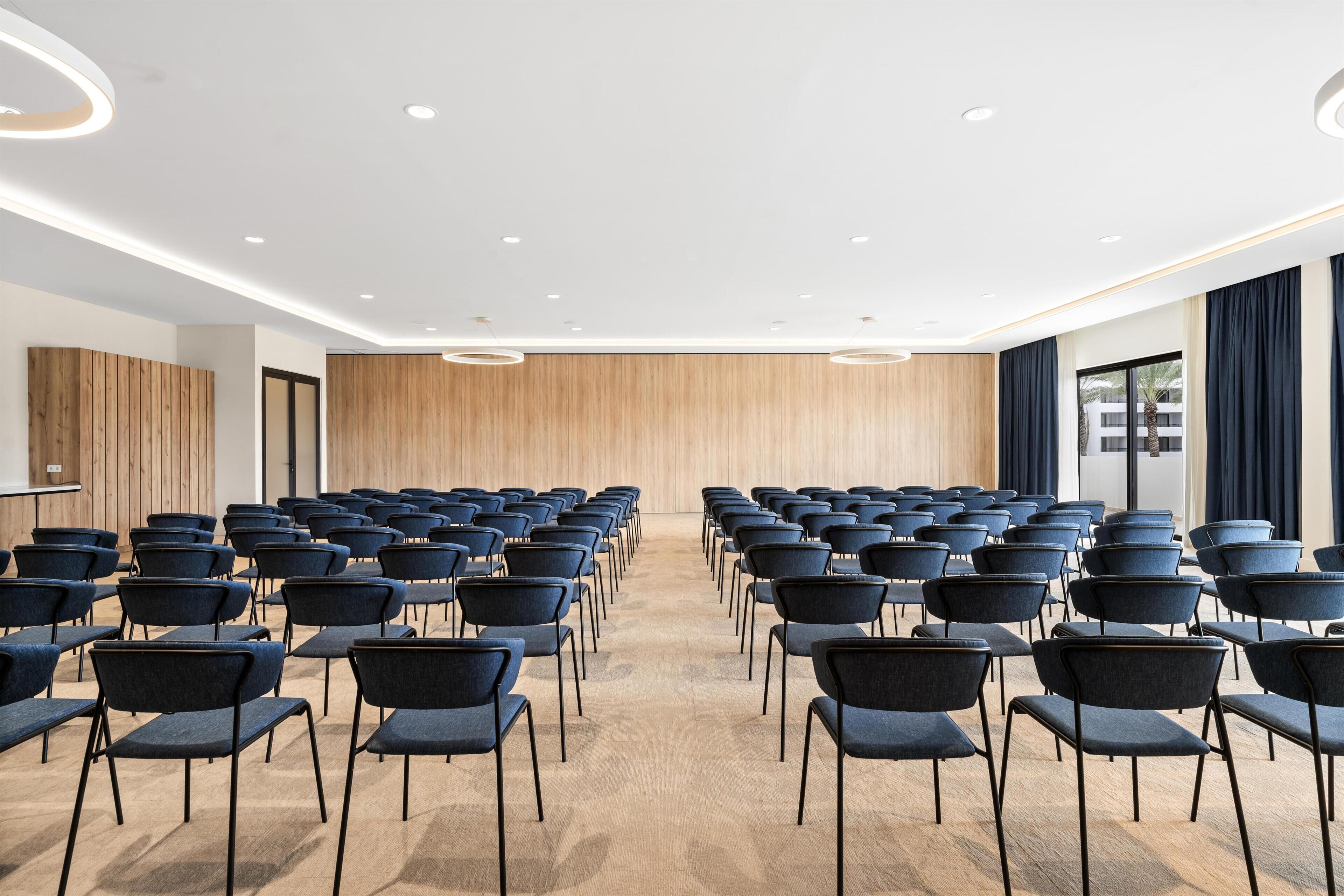 A conference room with rows of chairs facing a wooden panel wall, equipped with ceiling lights and a large window with curtains.