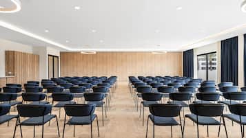 A conference room with rows of chairs facing a wooden panel wall, equipped with ceiling lights and a large window with curtains.