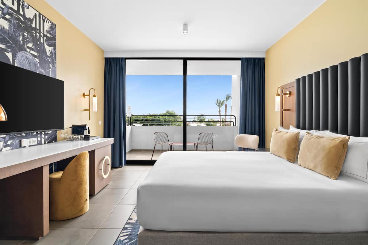 A hotel room with a large bed, a desk with a TV, a balcony with chairs, and a view of palm trees.