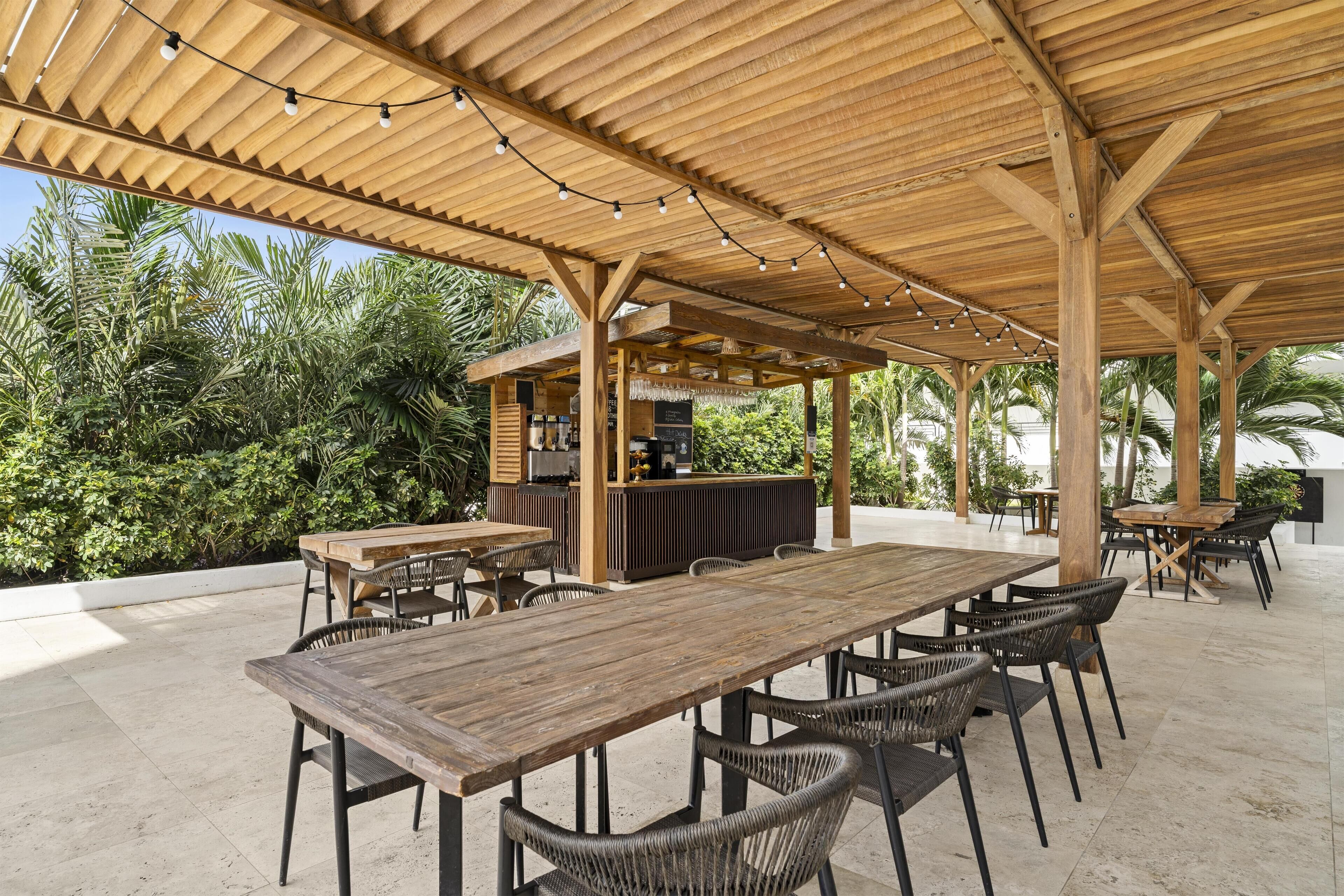 Eine Terrasse im Freien mit Holztischen und -stühlen, einer Bar und üppiger Bepflanzung im Hintergrund.