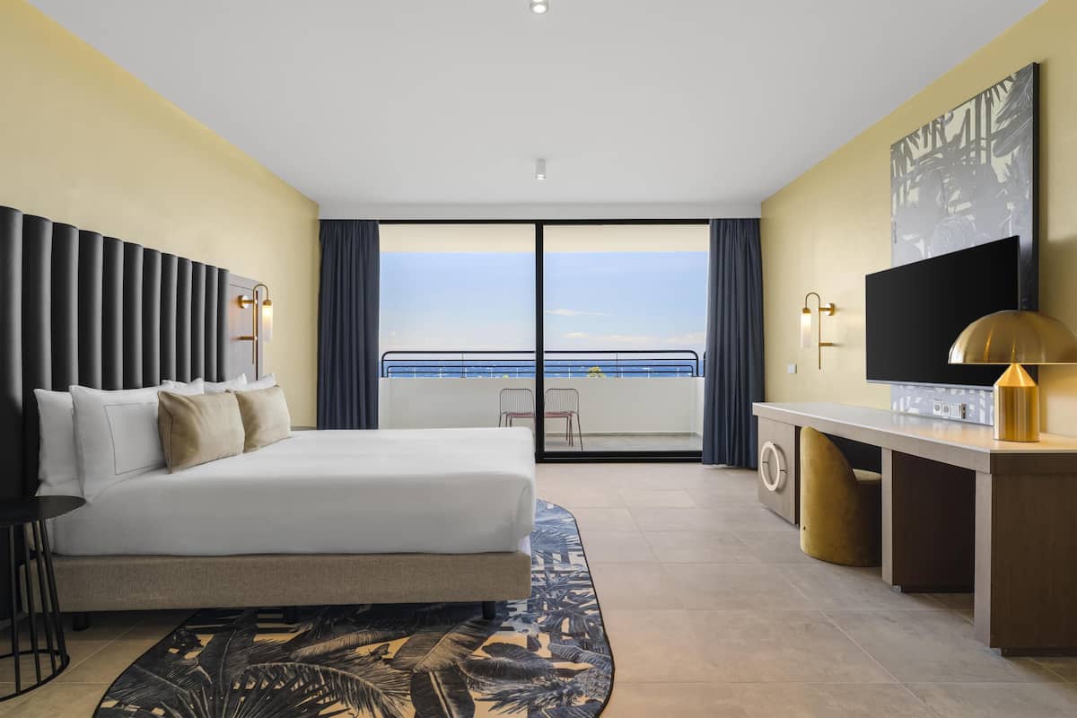 A hotel room with a bed, a desk, a television, and a view of the sea.