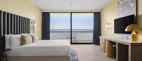 A hotel room with a bed, a desk, a television, and a view of the sea.