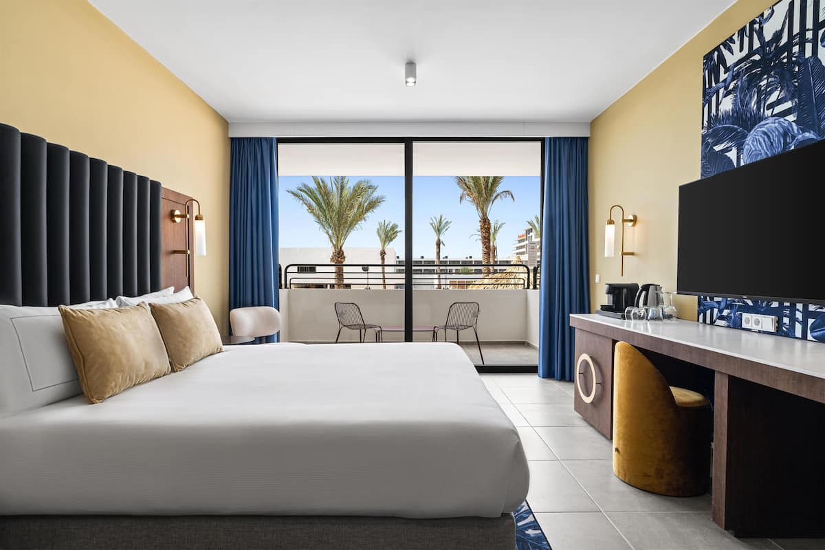 A hotel room with a large bed, a desk with a chair, a TV, and a balcony with palm trees and a view of the city.