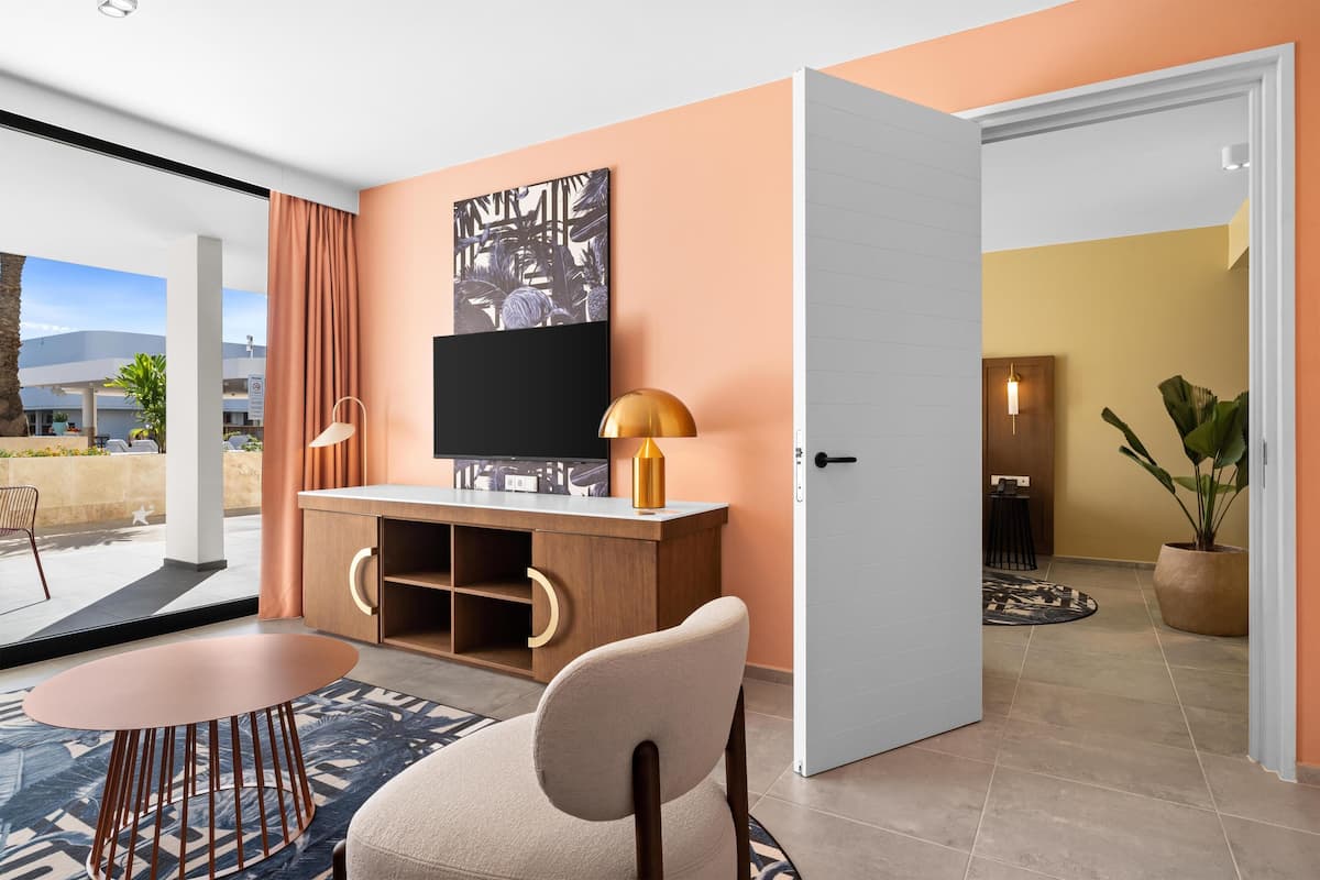 A modern hotel room with a large window, a flat-screen TV, a wooden desk, a round table, and a comfortable chair.