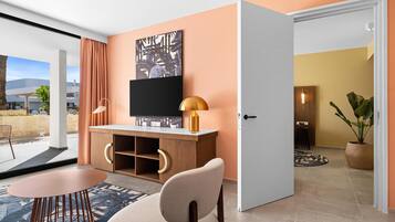 A modern hotel room with a large window, a flat-screen TV, a wooden desk, a round table, and a comfortable chair.