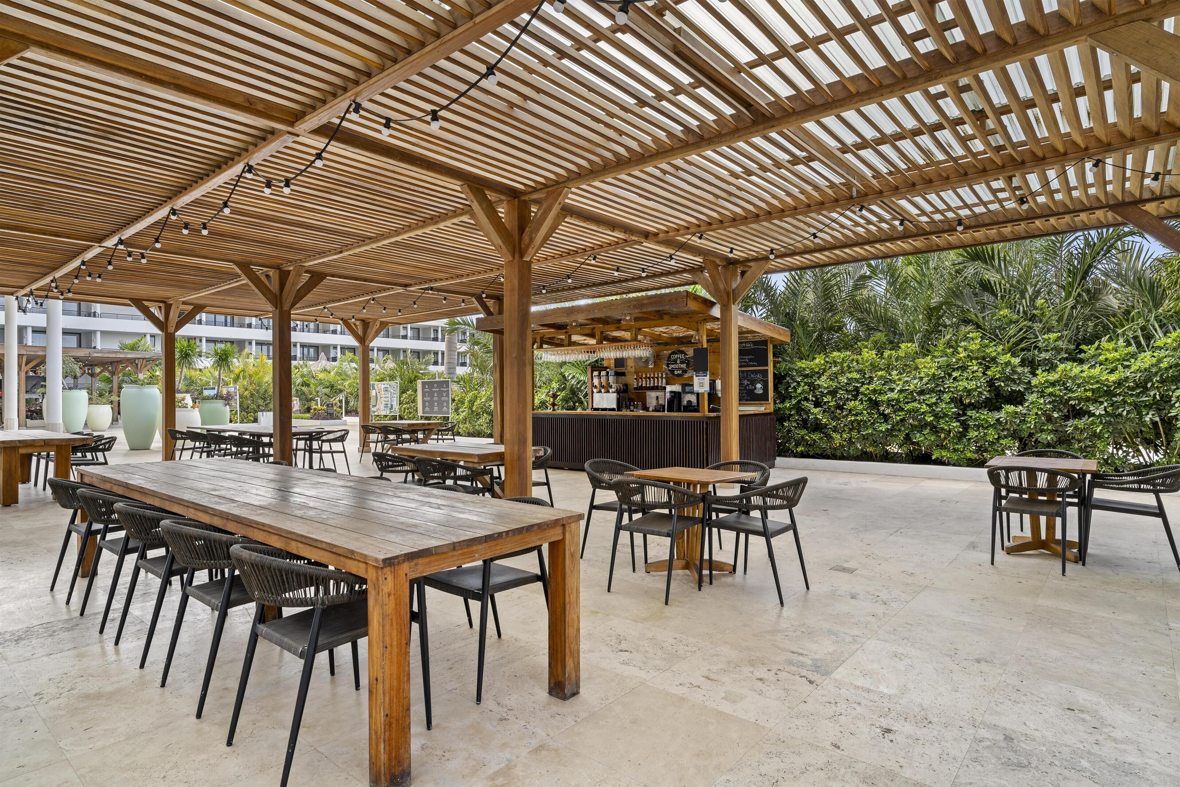 An outdoor patio area with wooden tables and chairs, a bar area, and a view of greenery and buildings in the background.