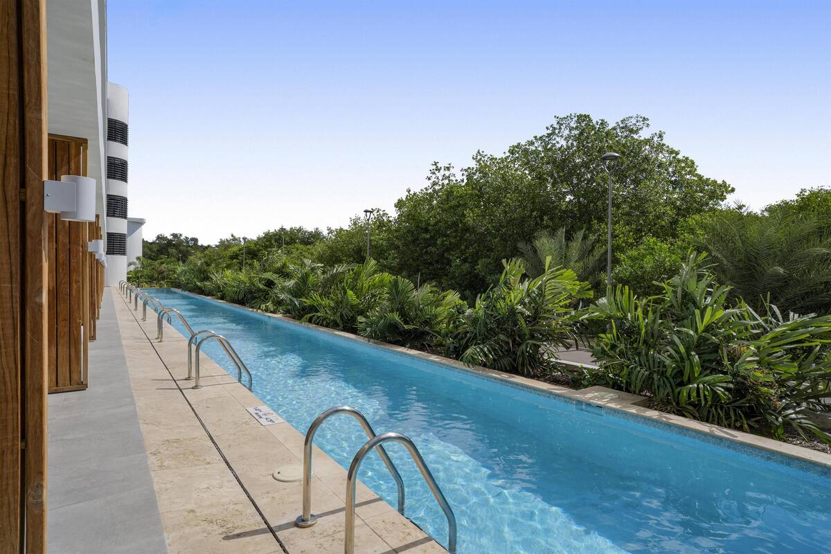 A swimming pool with metal railings, surrounded by lush greenery and a modern building with white and wooden elements.