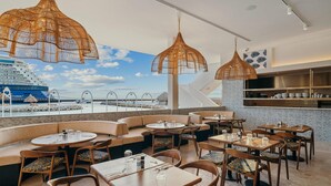 A restaurant with round tables and booths, large woven pendant lights, and a view of a cruise ship.