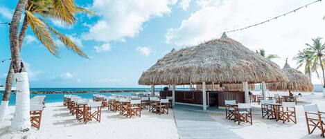 Un resort frente a la playa con estructuras de techo de paja, sillas de descanso blancas y una terraza de madera que da a la playa.