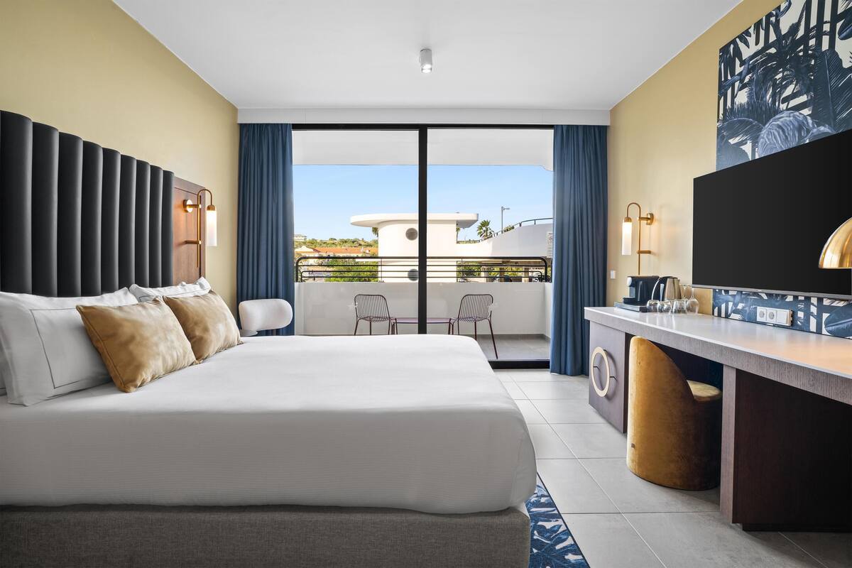 A modern hotel room with a large bed, a desk, and a balcony with chairs.