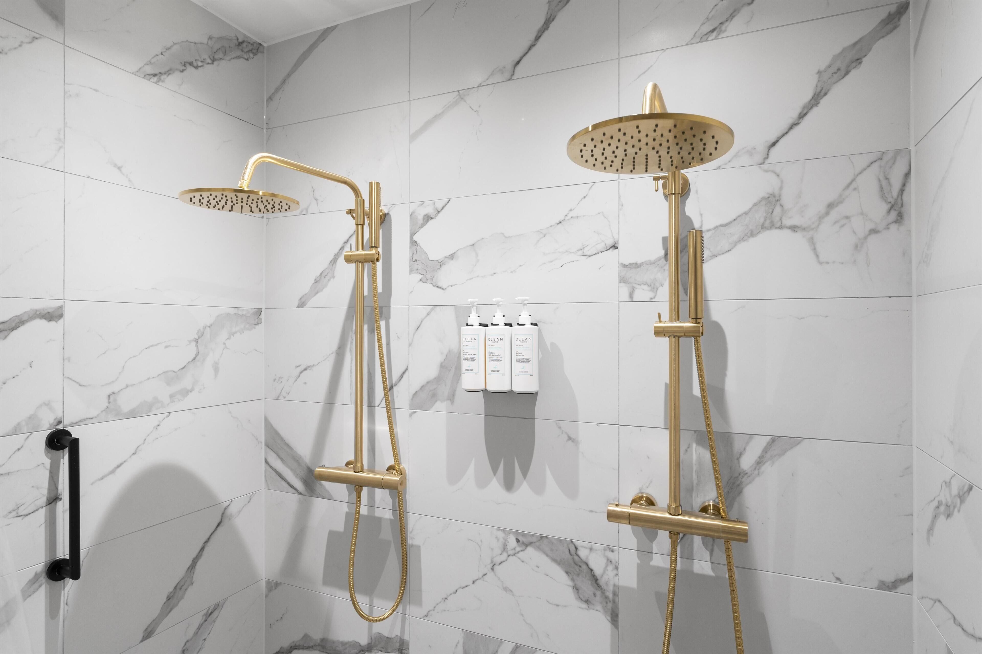 A bathroom with two gold showerheads and a black grab bar on the wall.