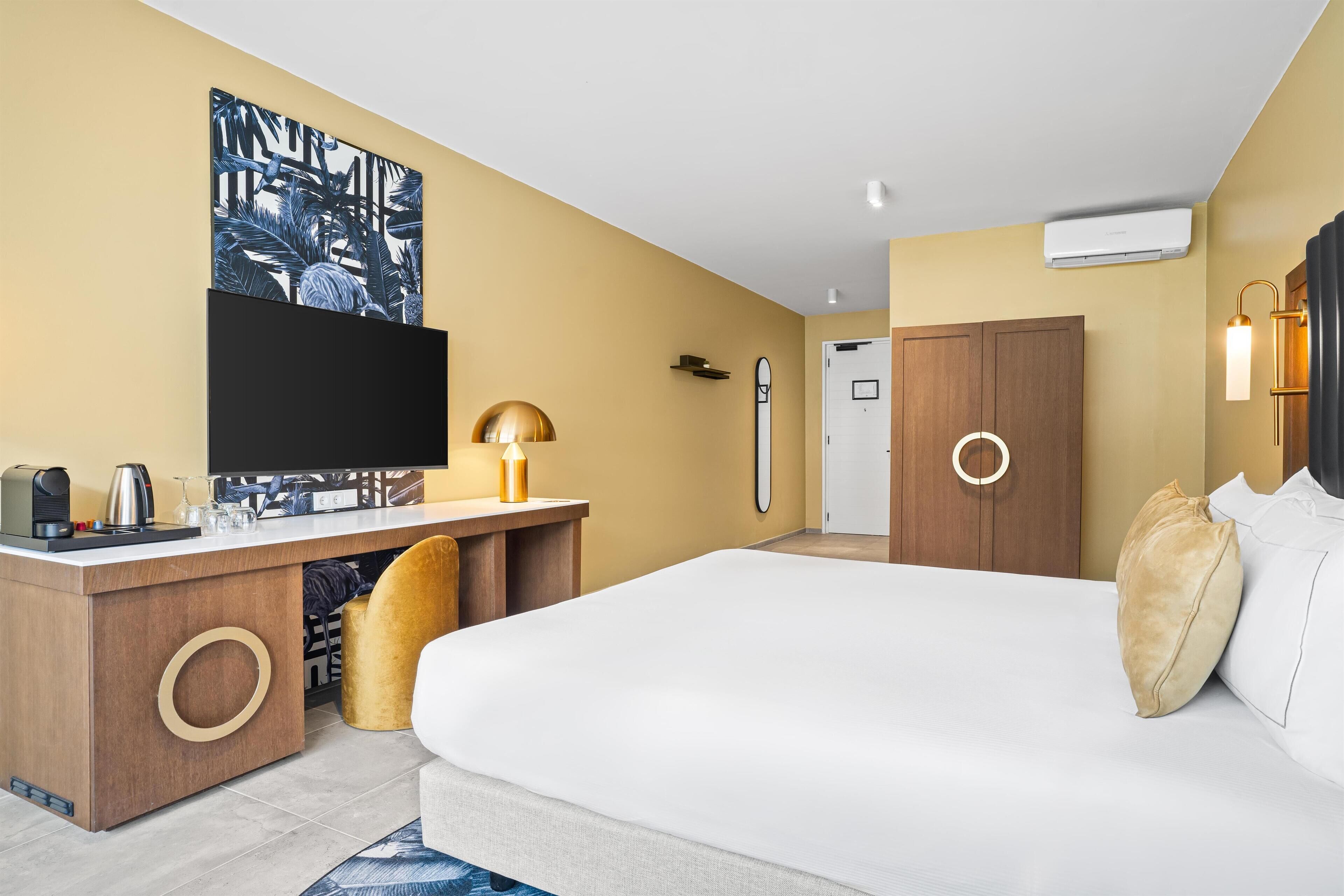 Ein Hotelzimmer mit einem großen Bett, einem Schreibtisch, einem Flachbildfernseher, einer Kaffeemaschine und einer dekorativen Lampe.
