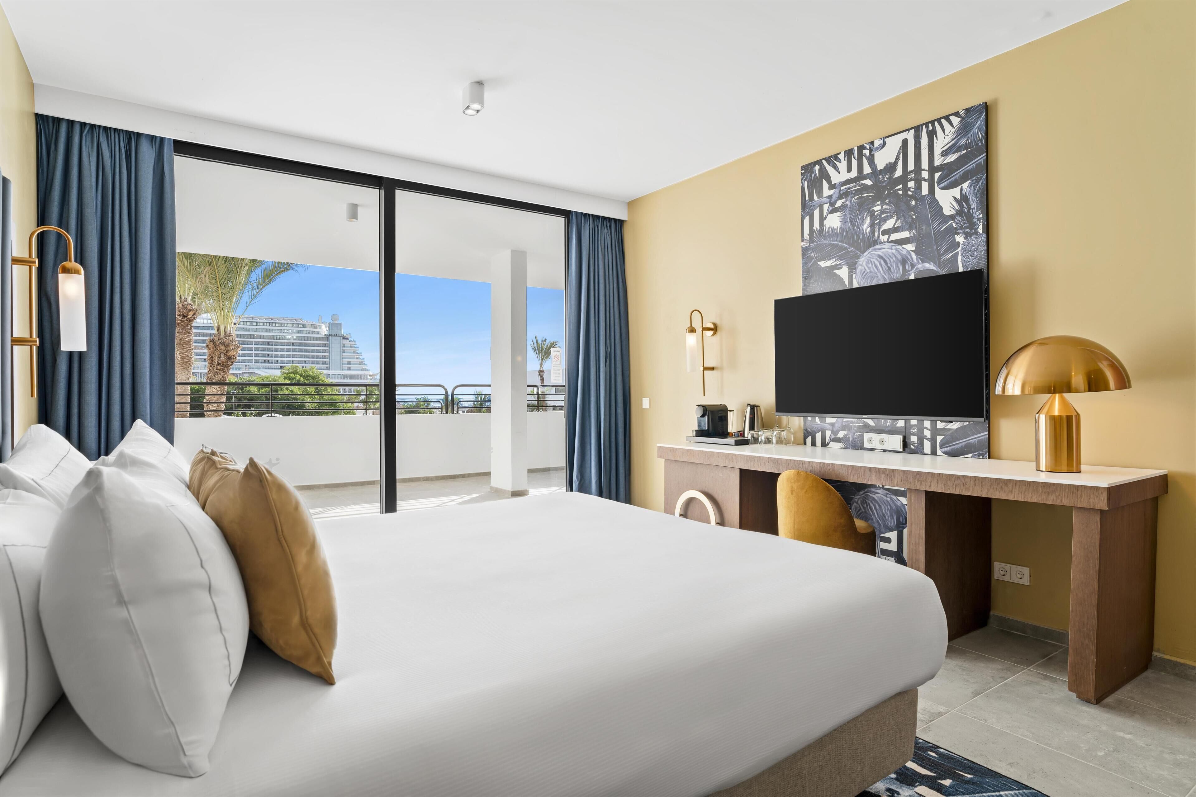 A hotel room with a large bed, a desk with a television, and a view of a building and palm trees.