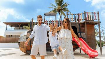 Una familia caminando en la playa, con una estructura de juegos de madera y un tobogán rojo al fondo.