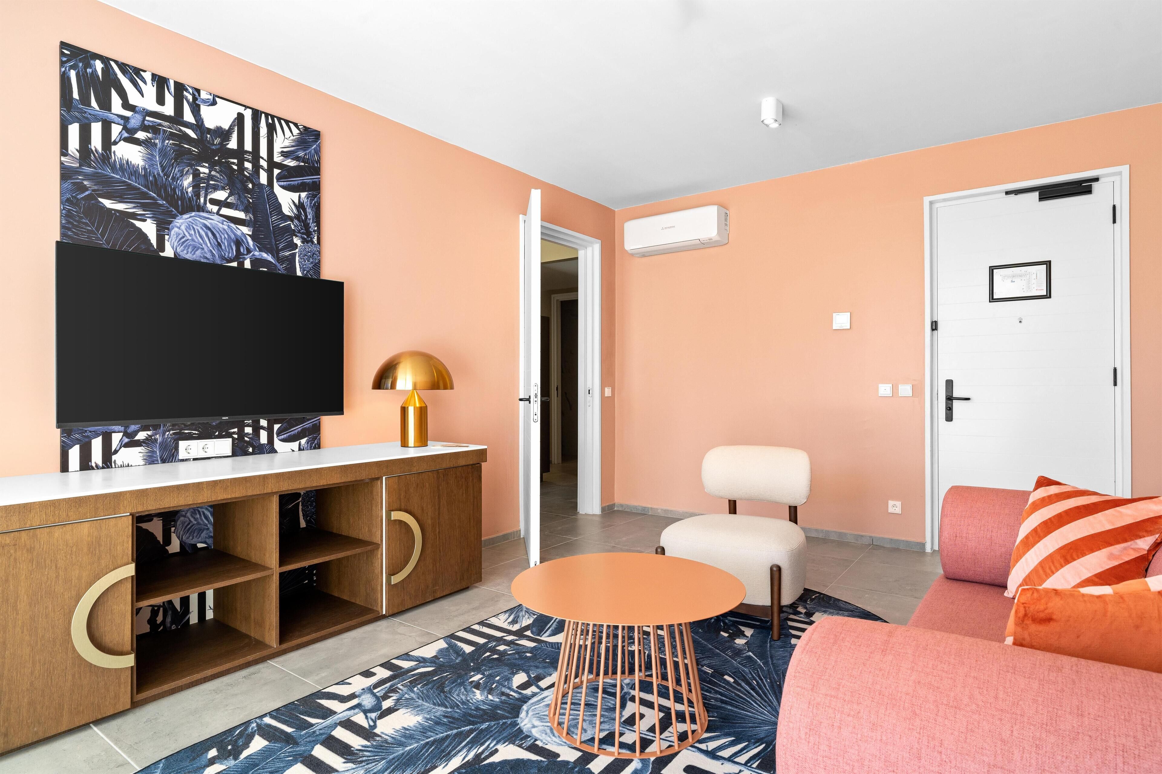 A living room with a flat-screen TV, a wooden cabinet, a round orange table, a sofa, and a door.