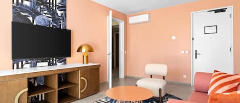 A living room with a flat-screen TV, a wooden cabinet, a round orange table, a sofa, and a door.
