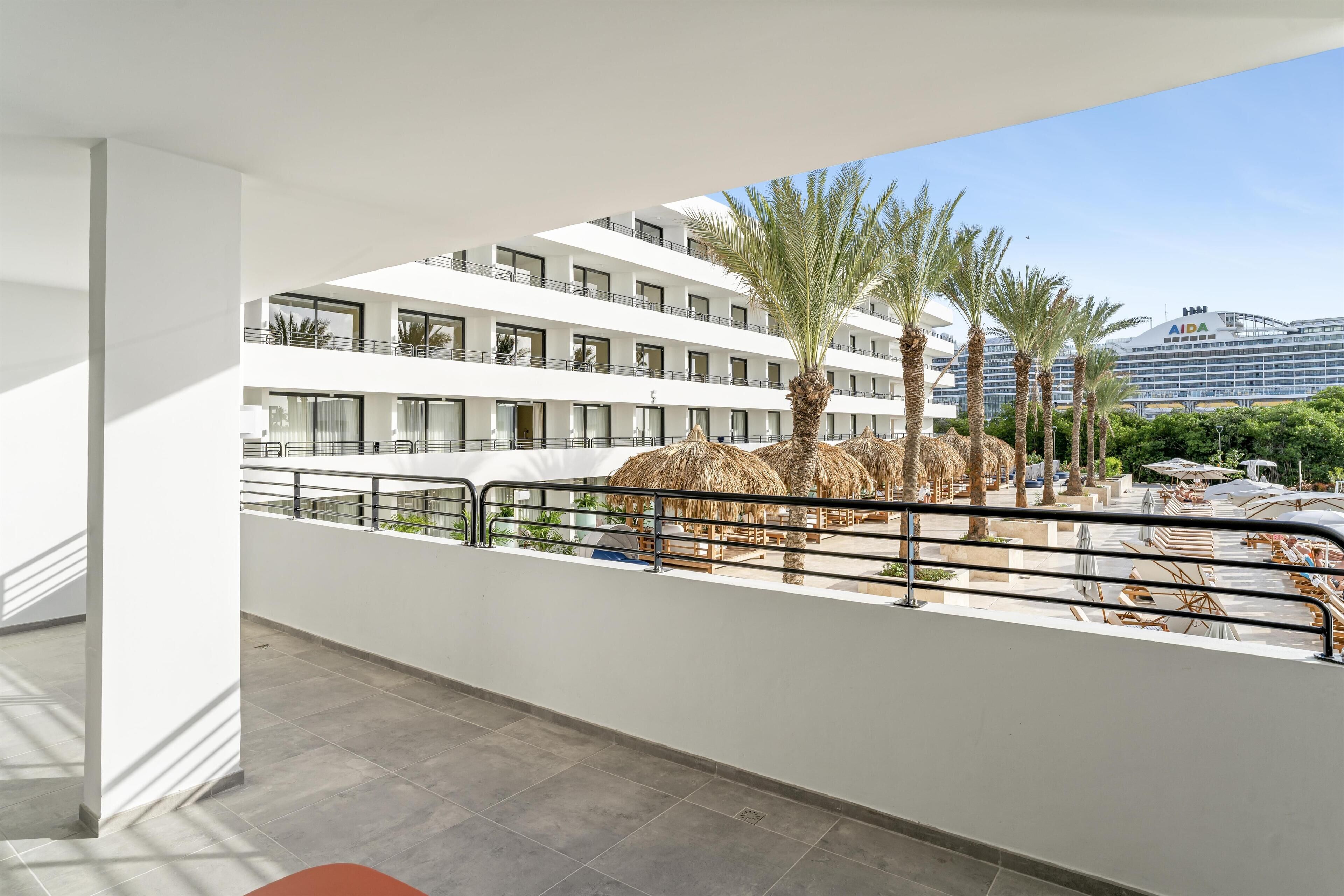 A hotel balcony with a view of a pool area, palm trees, and a building with 'AIDA' signage.