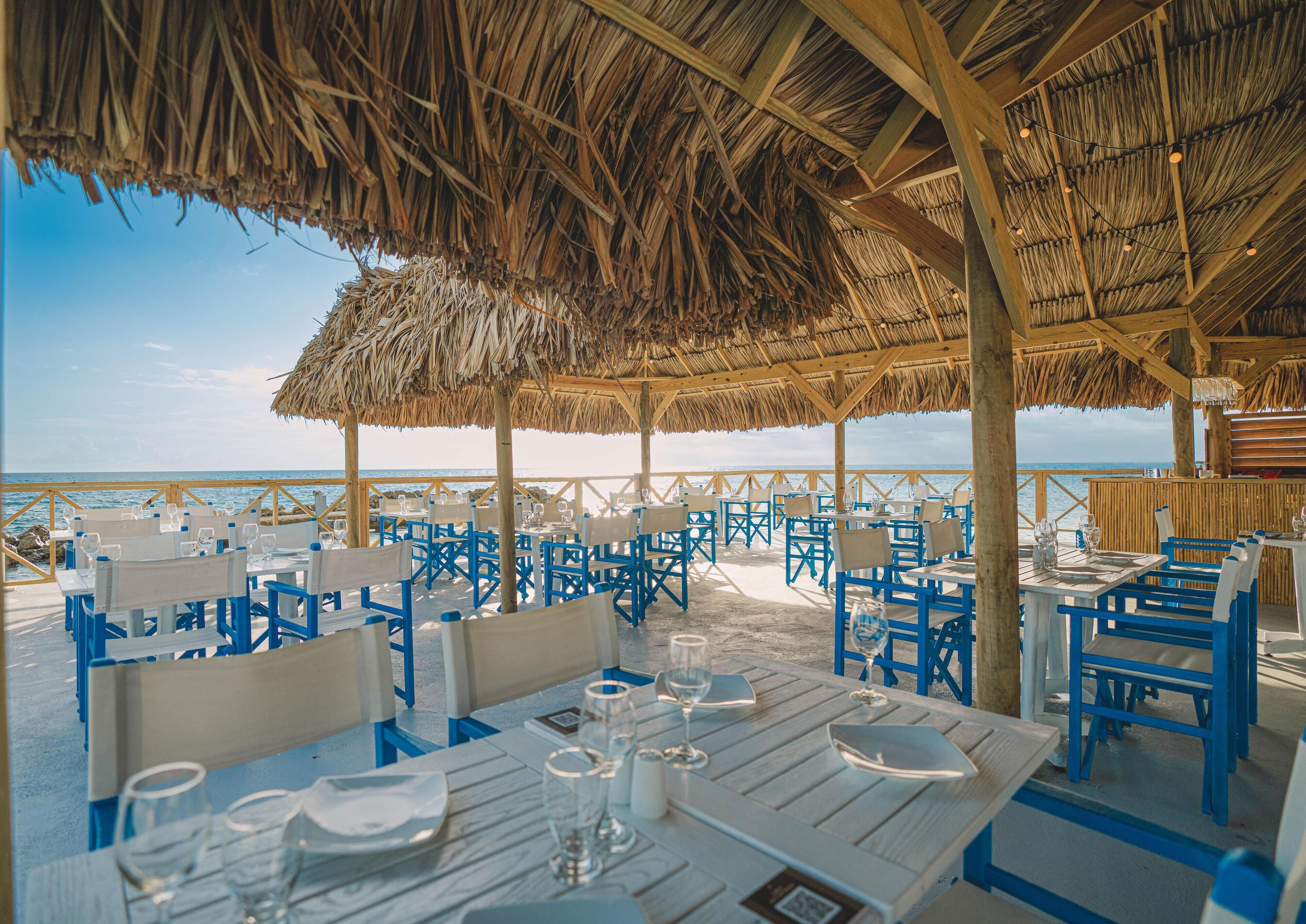 An outdoor dining area with tables and chairs, a thatched roof, and a view of the ocean.