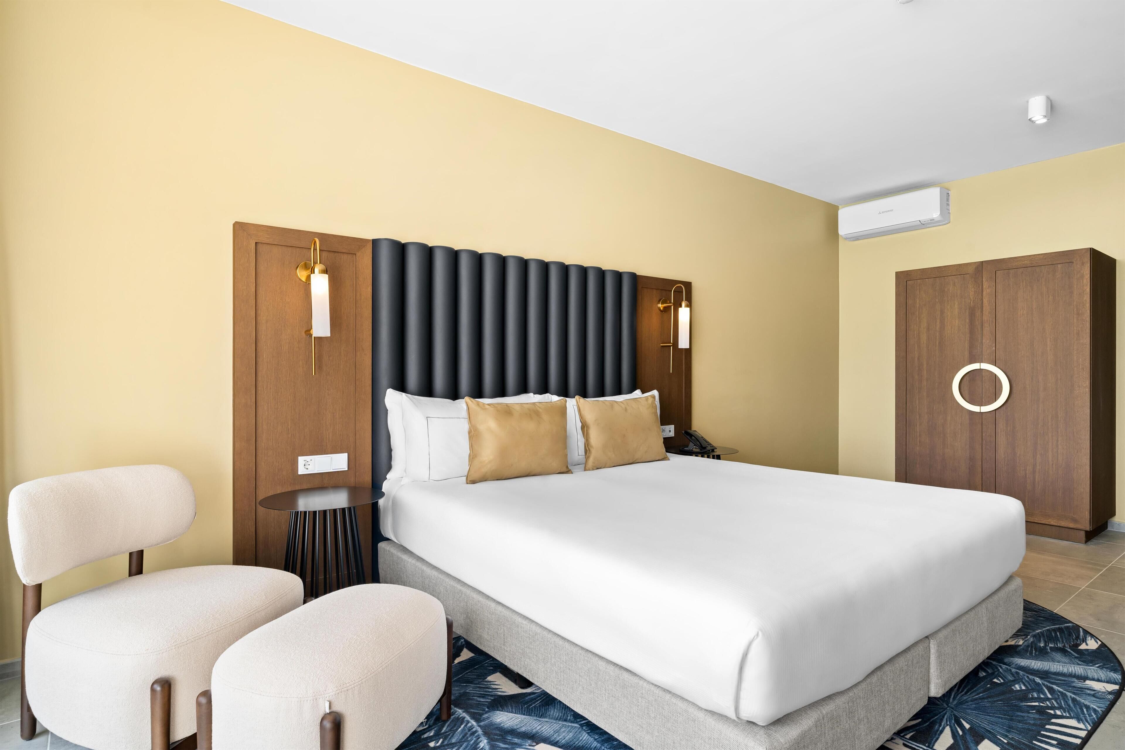 Quarto de hotel com uma cama grande, duas cadeiras, uma mesinha e um guarda-roupa.