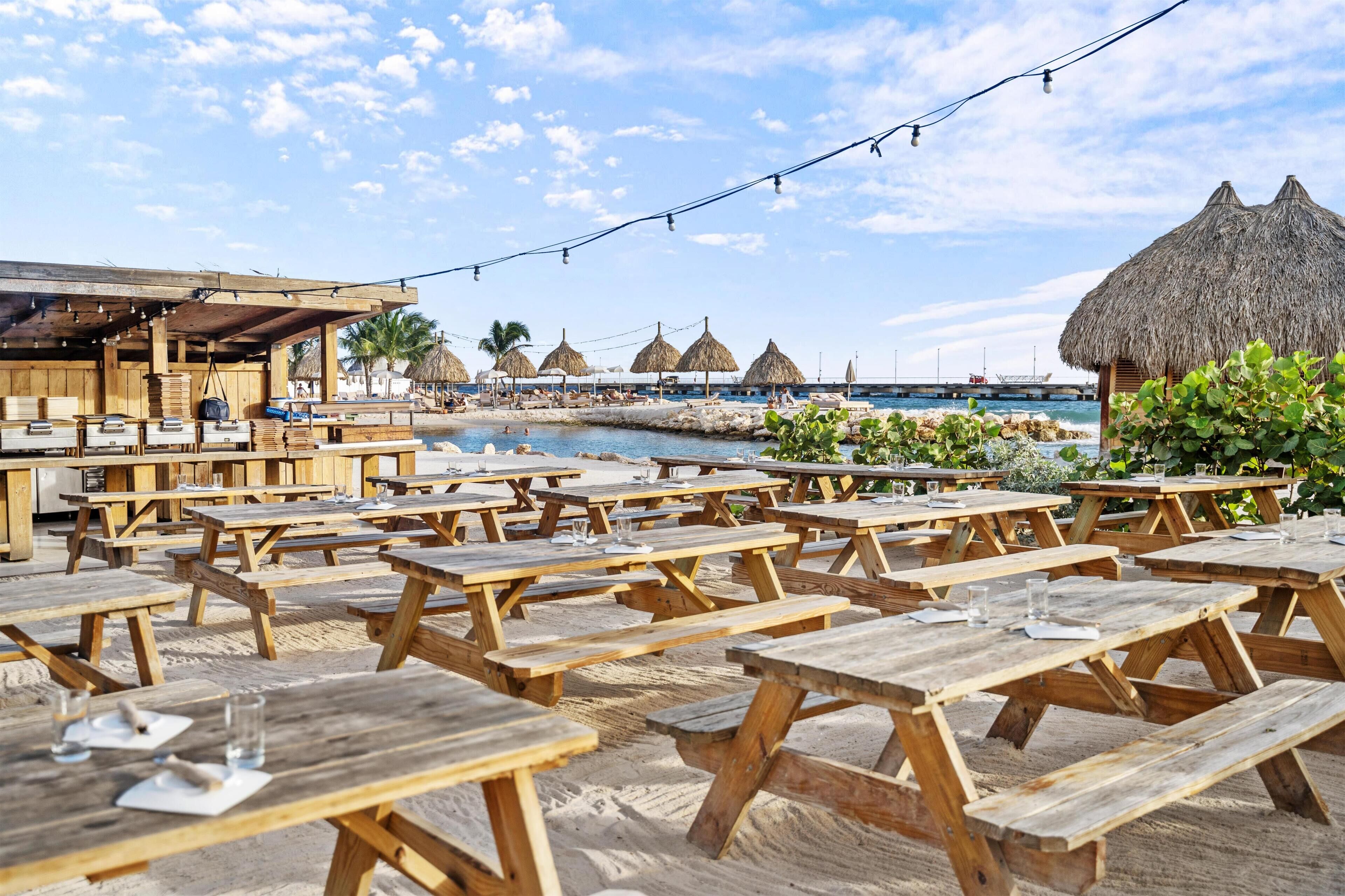 Un espace de restauration en plein air, avec des tables de pique-nique en bois et des parasols en chaume, offrant une vue sur une plage et l’océan.