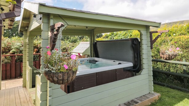 Ein Whirlpool unter einem grünen Pavillon mit einer hölzernen Schaukel, umgeben von üppigem Grün und einer Holzterrasse.