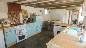 A kitchen with a stove, sink, and washing machine. There is a dining table and chairs in the background. - Brewers Cottage (Umberleigh)