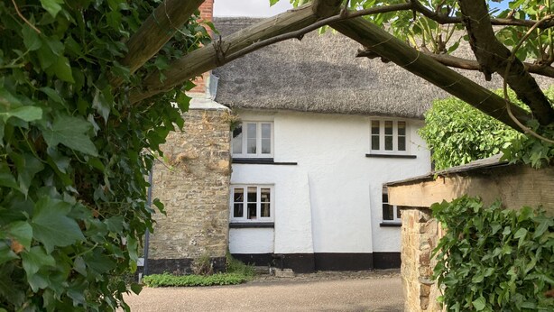 A white building with black trim and a thatched roof, surrounded by greenery and cobblestone paths. - Brewers Cottage (Umberleigh)