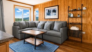 A modern living room with a grey sectional sofa, a wooden coffee table, and a blue patterned rug. There are two windows with curtains, a ceiling fan, and a wooden wall panel with a shelf. - ML 342: Breathtaking Views & Prime Location for the Perfect Summer Stay (Snowshoe)