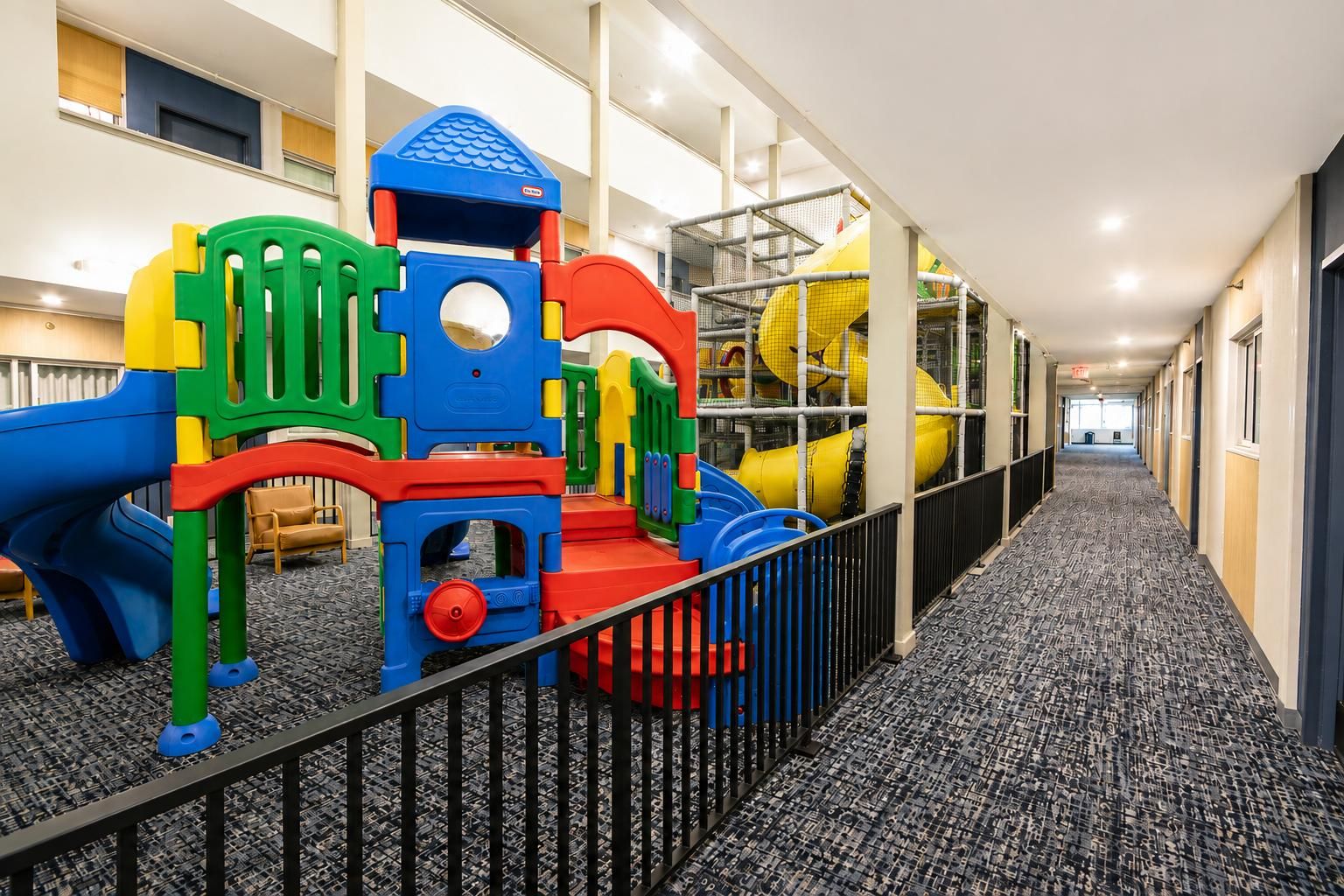 A play area with colorful play structures and a slide, located in a hallway with patterned carpet and a metal railing.