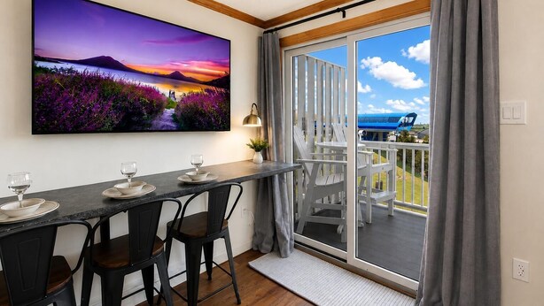 A dining area with a table set for two, a large painting of a sunset landscape, and a balcony with a view of the sky and a distant building. - ML 342: Breathtaking Views & Prime Location for the Perfect Summer Stay (Snowshoe)